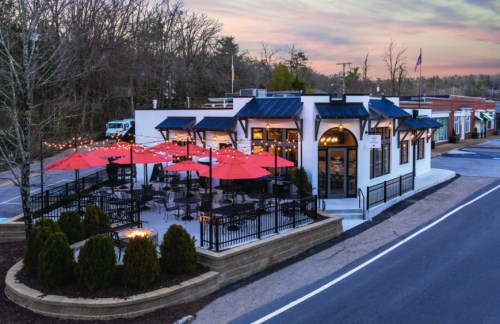 A modern restaurant with outdoor seating under red umbrellas, surrounded by trees and a sunset sky.