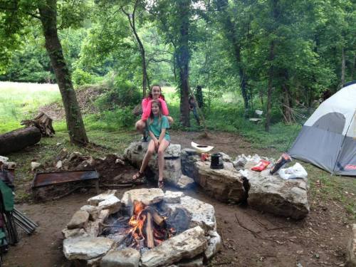 Two people sit by a campfire in a wooded area, with a tent and camping gear visible nearby.