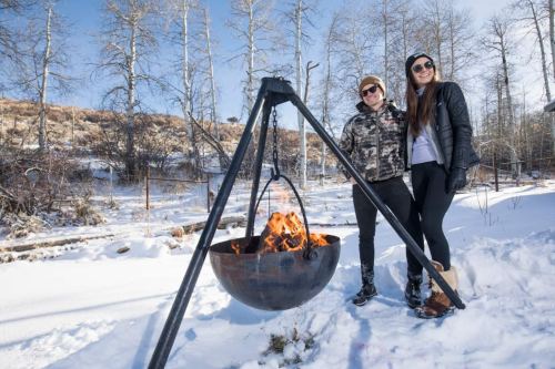 Two people stand beside a fire pit in a snowy landscape, smiling and dressed warmly in winter attire.