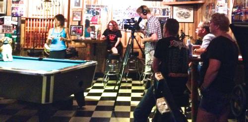 A group of people filming in a bar with a pool table, featuring a woman with a camera and others watching.