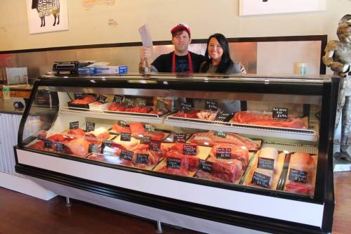 A butcher and a woman stand behind a display case filled with various cuts of meat in a shop.