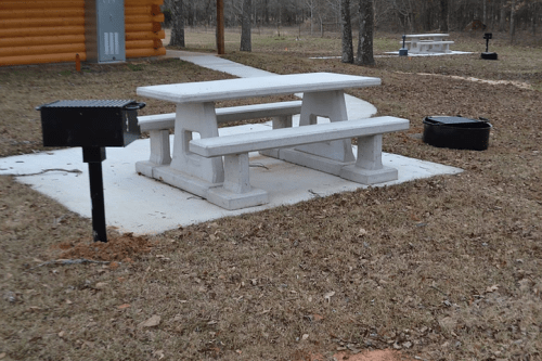 A concrete picnic table with a grill and fire pit on a grassy area near a cabin in a wooded setting.