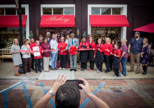 A group of people, including bakery staff and guests, celebrate a ribbon-cutting ceremony outside a bakery.