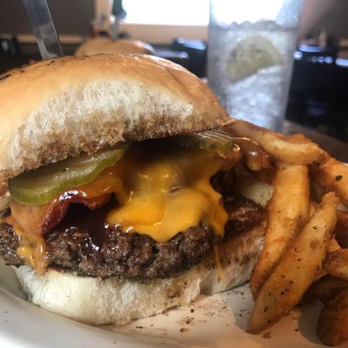 A juicy cheeseburger with bacon, pickles, and melted cheese, served with crispy fries and a drink in the background.
