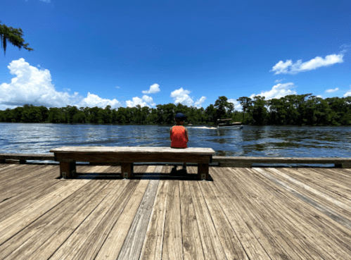 A child sits on a wooden bench by a river, gazing at the water under a bright blue sky with fluffy clouds.