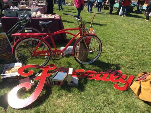 A vintage red bicycle next to a large, colorful "Friendly" sign on grass, with people browsing in the background.