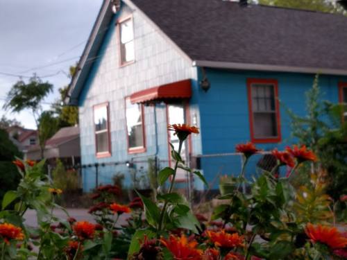 A blue house with orange trim and flowers in the foreground, set against a cloudy sky.