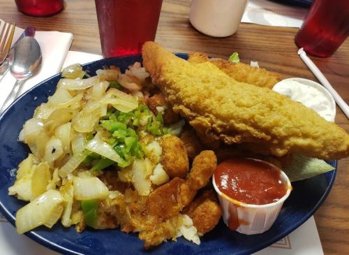 A plate of fried fish fillets with sautéed onions, green peppers, and a side of dipping sauce on a wooden table.