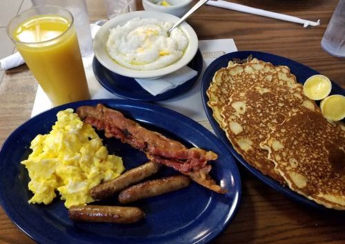 A breakfast plate with scrambled eggs, bacon, sausage, pancakes, and a glass of orange juice.