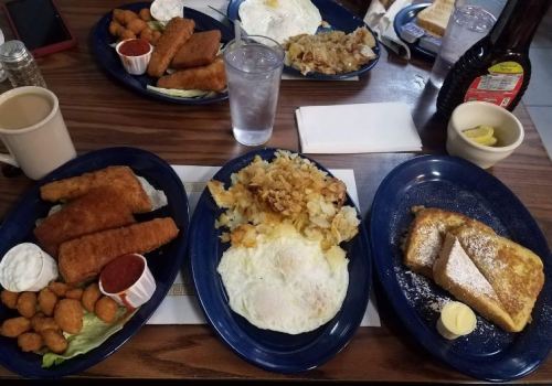A table with three plates of breakfast: eggs, French toast, hash browns, fish sticks, and dipping sauces, with drinks.
