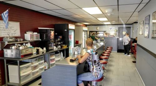 A cozy diner interior with a counter, red stools, and patrons enjoying their meals. Walls adorned with framed pictures.