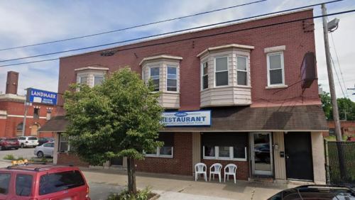 A brick building with a restaurant sign, featuring large windows and a tree in front, parked cars nearby.