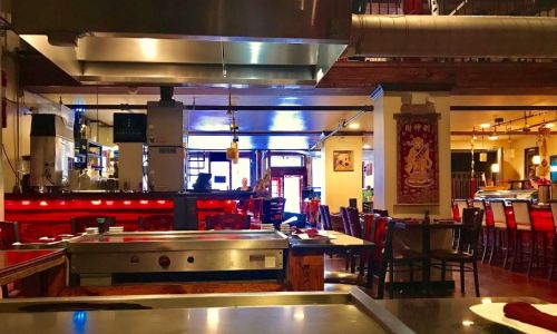 Interior of a restaurant featuring a grill, red lighting, wooden tables, and a bar area in the background.