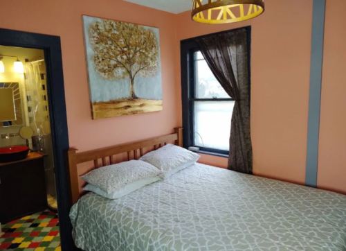 Cozy bedroom with a wooden bed, colorful rug, and a tree painting on the wall. A bathroom is visible in the background.