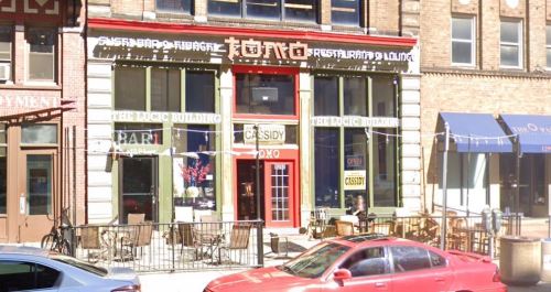 Exterior of a restaurant with a red entrance, outdoor seating, and large windows, located in a brick building.
