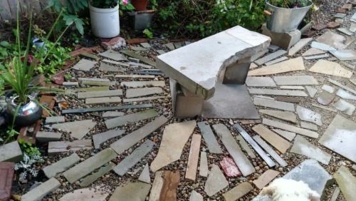 A stone bench surrounded by a circular pattern of various stones and gravel, with plants in pots nearby.