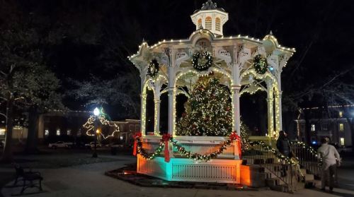 A beautifully lit gazebo adorned with Christmas decorations and a large tree, set in a park at night.