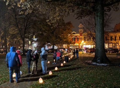 A nighttime scene of people walking along a park path lined with glowing lanterns, surrounded by trees and buildings.