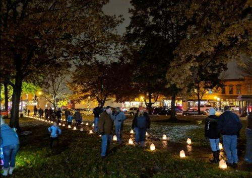 A nighttime scene of people walking along a path lined with glowing lanterns, surrounded by trees and buildings.