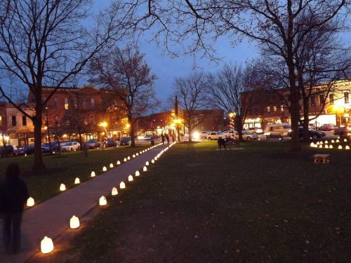 A park path lined with glowing lanterns at dusk, with trees and buildings in the background.