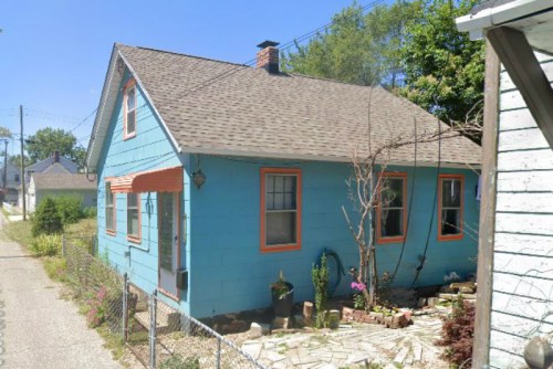 A small blue house with orange trim, surrounded by greenery and a gravel path, viewed from an angle.