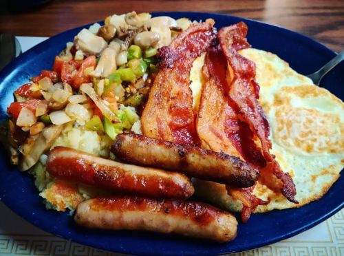 A hearty breakfast plate with bacon, sausages, fried egg, and a mix of sautéed vegetables and mashed potatoes.