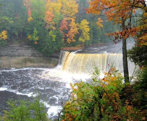 A waterfall cascades over rocky cliffs, surrounded by vibrant autumn foliage and misty air.
