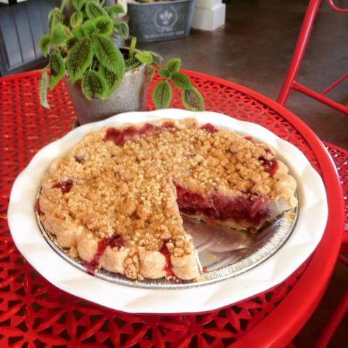 A cherry pie with a crumb topping sits on a red table, with a slice removed and a potted plant in the background.