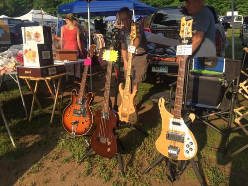 A variety of bass guitars displayed at an outdoor market, with people browsing in the background.