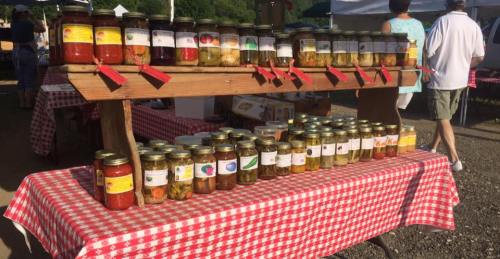 A wooden table displays various jars of colorful preserves and pickles, set against a checkered tablecloth at a market.