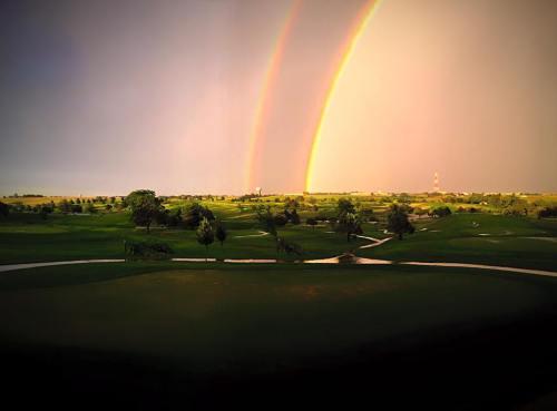 A vibrant landscape featuring a double rainbow over a lush green golf course under a dramatic sky.