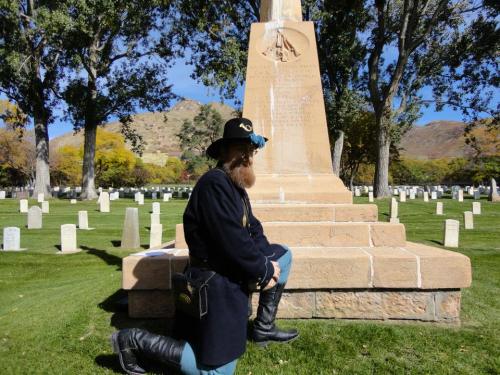 A person in historical military attire kneels by a monument in a cemetery, surrounded by gravestones and trees.
