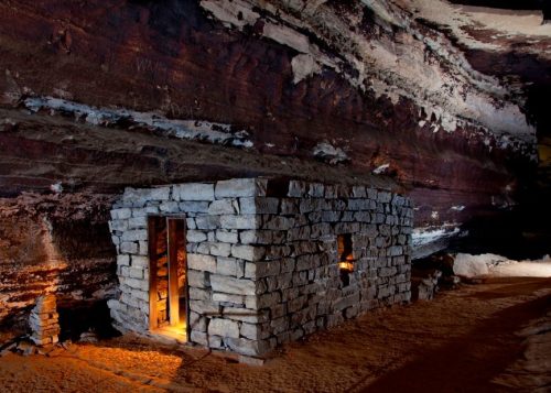 A small stone structure illuminated inside a cavern with layered rock walls and sandy ground.