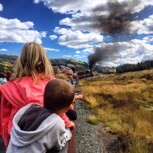 A group of people, including children, watch a steam train billowing smoke against a scenic landscape and blue sky.