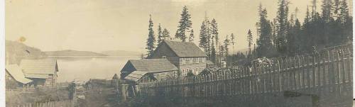 Black and white photo of a lakeside settlement with wooden buildings and trees, reflecting a serene landscape.