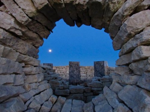 View through a stone archway, revealing a clear blue sky and a bright moon above ancient stone structures.
