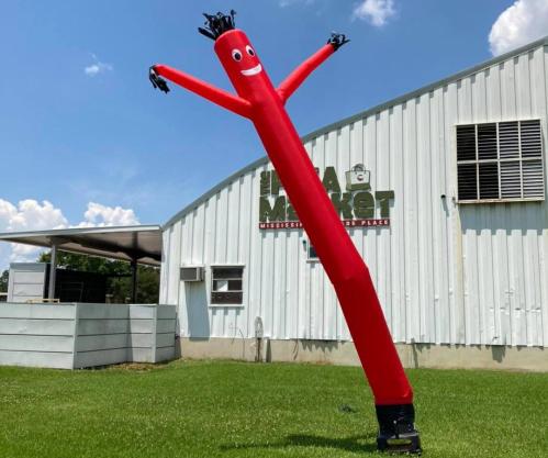 A tall, red inflatable tube man with a smiling face waves in front of a market building on a sunny day.