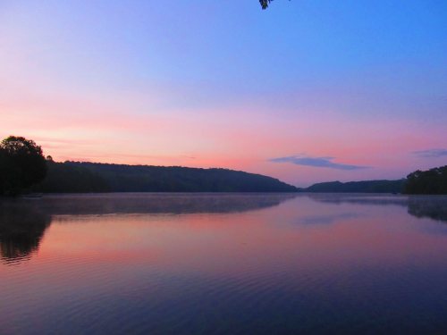 A serene lake at sunset, reflecting soft pink and blue hues with trees lining the shore.