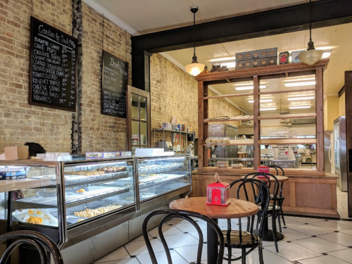 A cozy bakery interior with display cases of pastries, wooden counters, and black chairs in a brick-walled setting.