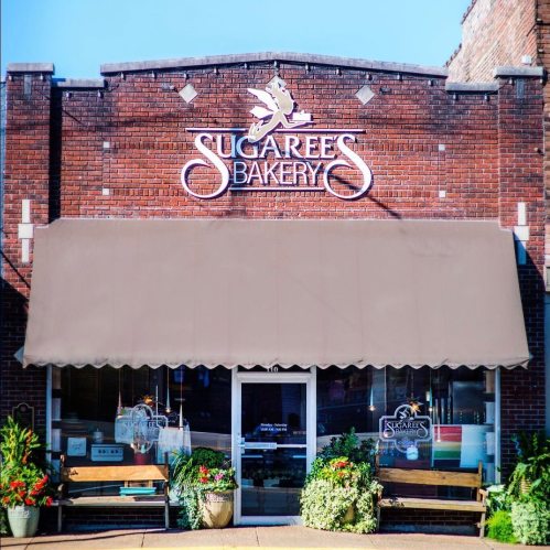 A brick bakery storefront with a brown awning and colorful flower planters, featuring the sign "Sugaree's Bakery."