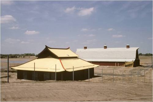 A yellow-roofed building and a red barn sit on a sandy landscape under a blue sky with scattered clouds.