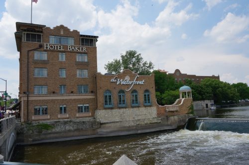 Historic Hotel Baker alongside the waterfront, with a dam and lush greenery under a partly cloudy sky.