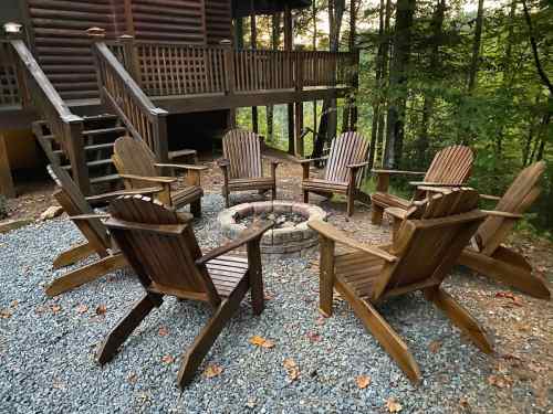 A circle of wooden Adirondack chairs around a stone fire pit, surrounded by trees and a log cabin in the background.