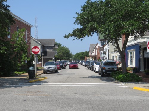 A quiet street lined with parked cars, featuring trees and buildings on either side under a clear blue sky.
