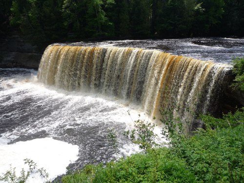 A cascading waterfall flows over a rocky ledge, surrounded by lush greenery and a dark river below.