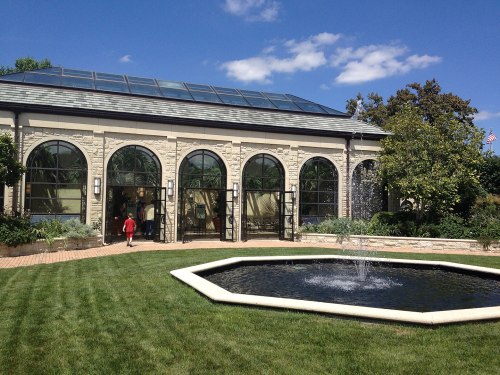 A modern building with arched windows, surrounded by greenery and a fountain in a landscaped garden under a blue sky.