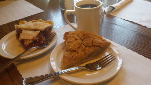 Two slices of pie on white plates, one cherry and one apple crumb, beside a cup of coffee on a wooden table.