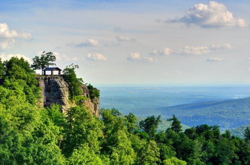 A scenic view of a cliffside house surrounded by lush greenery and distant mountains under a blue sky with clouds.