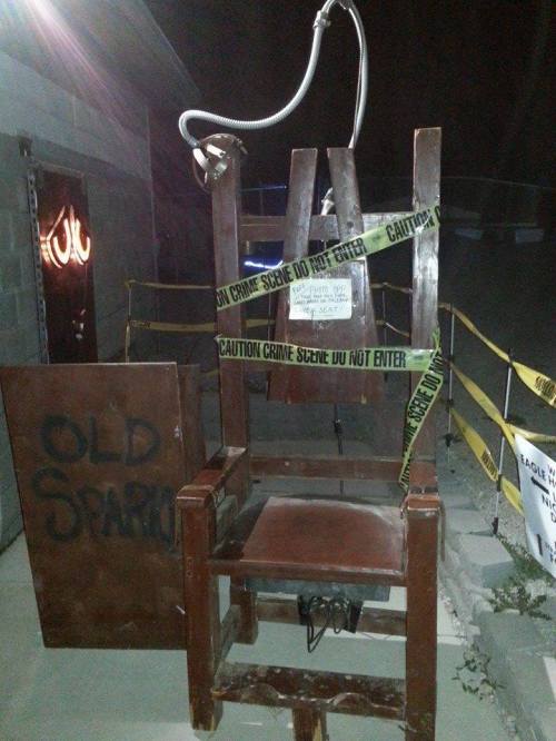 A wooden electric chair with caution tape, set in a dimly lit outdoor area, suggesting a crime scene.