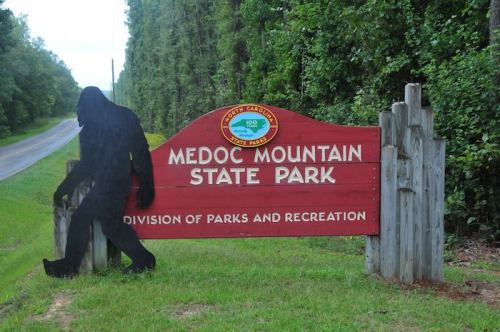Sign for Medoc Mountain State Park with a silhouette of a Bigfoot figure beside it, surrounded by trees and a road.